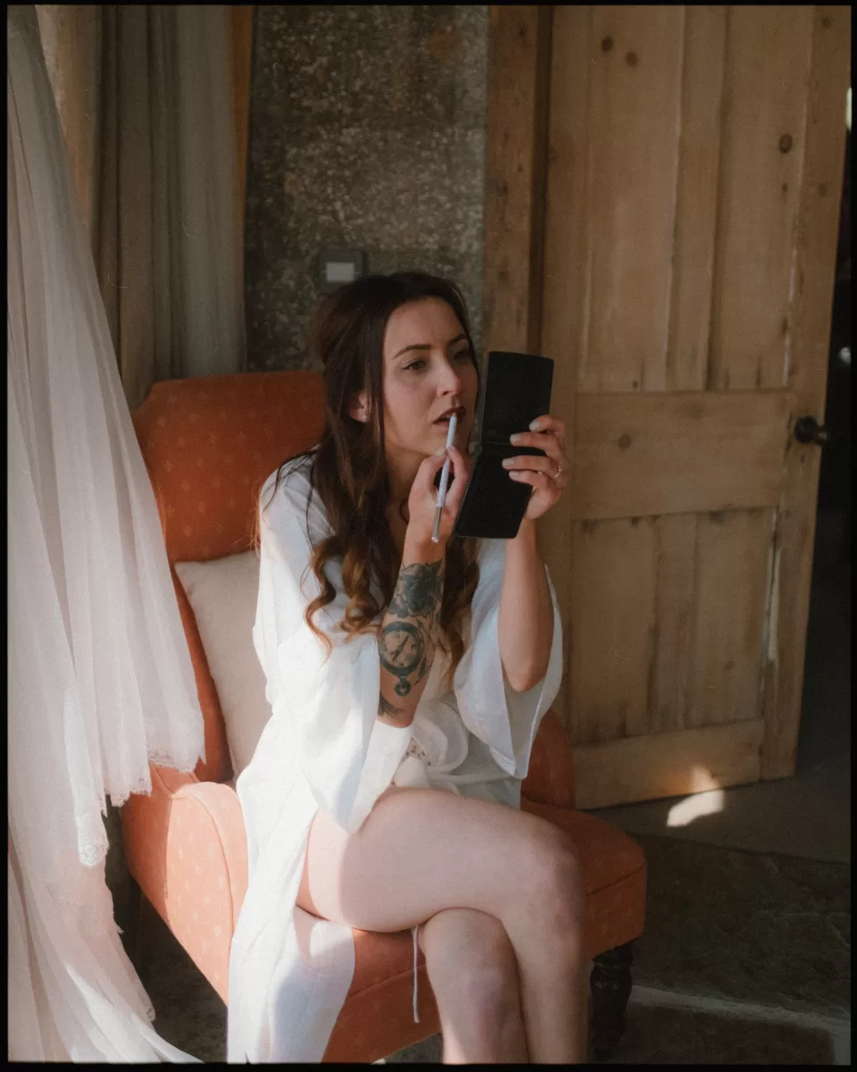 bride doing her own makeup with a pocket mirror next to wedding dress